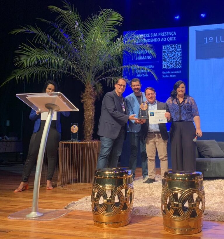 Registro de 4 pessoas segurando o prêmio Ser Humano. Um coqueiro ao lado esquerdo e uma tela azul com qr code ao fundo.