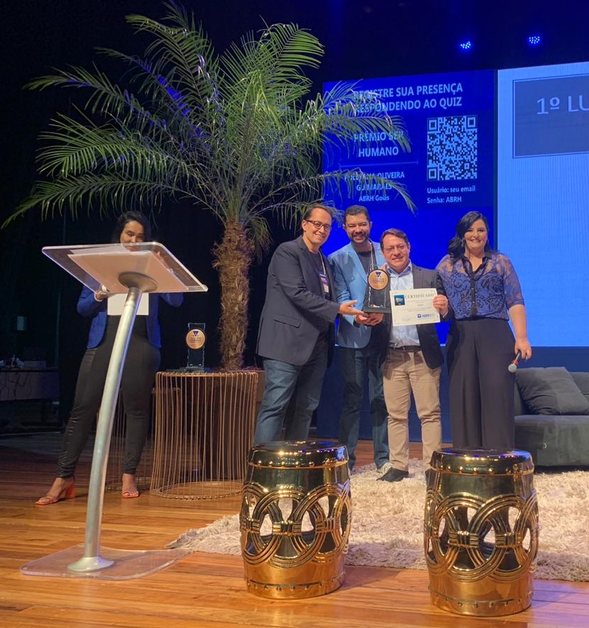 Registro de 4 pessoas segurando o prêmio Ser Humano. Um coqueiro ao lado esquerdo e uma tela azul com qr code ao fundo.